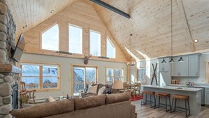 A spacious living area with a high ceiling, large windows, a stone fireplace, and a kitchen with bar stools.