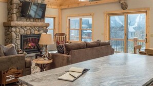 A cozy living room with a stone fireplace, a large sofa, a dining table, and a TV mounted on the wall.
