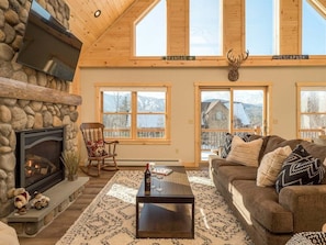 A cozy living room with a stone fireplace, a wooden ceiling, and large windows.
