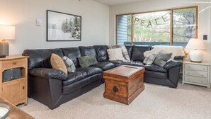A living room with a black leather sofa, a wooden coffee table, a side table, and a lamp. - Brookside II B-107 - Sunday River Slopeside Condominium (Newry)
