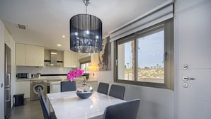 A modern kitchen with white cabinets, a dining table with chairs, a washing machine, and a large window with a view of palm trees. - Apartamento Vistabella (Vistabella Golf) (Orihuela Costa)