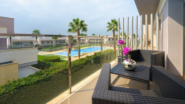 A balcony with wicker furniture, a glass railing, and a view of a swimming pool and palm trees. - Apartamento Vistabella (Vistabella Golf) (Orihuela Costa)