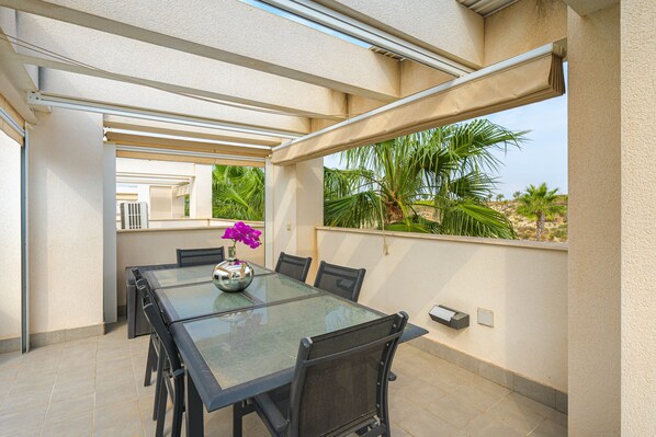 A covered outdoor dining area with a glass table and chairs, a decorative vase, and a view of palm trees and a building in the background.