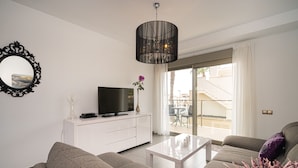 A modern living room with a flat-screen TV on a white cabinet, a grey sofa, a coffee table, and a large window with a view of a balcony. - Apartamento Vistabella (Vistabella Golf) (Orihuela Costa)