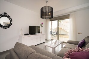 En moderne stue med et fladskærms-tv på et hvidt skab, en grå sofa, et sofabord og et stort vindue med udsigt til en balkon.