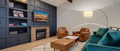 A modern living room with a fireplace, leather armchairs, a teal sofa, and a mounted TV.