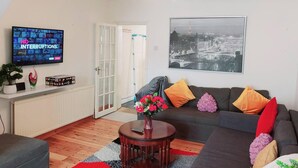 A living room with a television displaying 'NO INTERRUPTIONS', a sofa with colorful cushions, a round coffee table with a vase of flowers, and a black and white cityscape painting on the wall. - Beautiful 3-bed House in Chatham (Chatham)