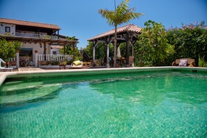Une piscine aux eaux cristallines, dotée d’une terrasse en bois et de chaises longues, entourée d’une végétation luxuriante et bordée d’un bâtiment à deux étages avec un balcon.