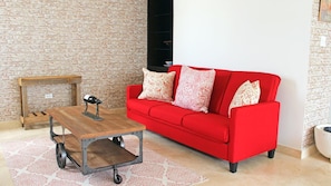 A living room with a red sofa, patterned cushions, a wooden coffee table with wheels, a small wooden side table, and a patterned rug.