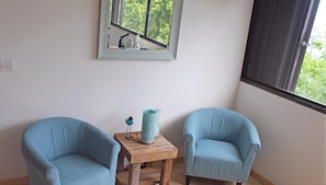 A room with two light blue armchairs, a small wooden table with a blue vase, and a window with a view of greenery.