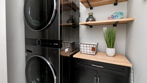 Un espace buanderie compact comprenant deux machines à laver superposées, une étagère en bois agrémentée d’objets décoratifs et une plante en pot.