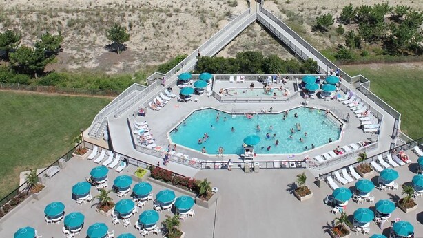 Vue aérienne d’un complexe hôtelier avec une piscine centrale entourée de transats et de parasols, et un paysage verdoyant en arrière-plan.