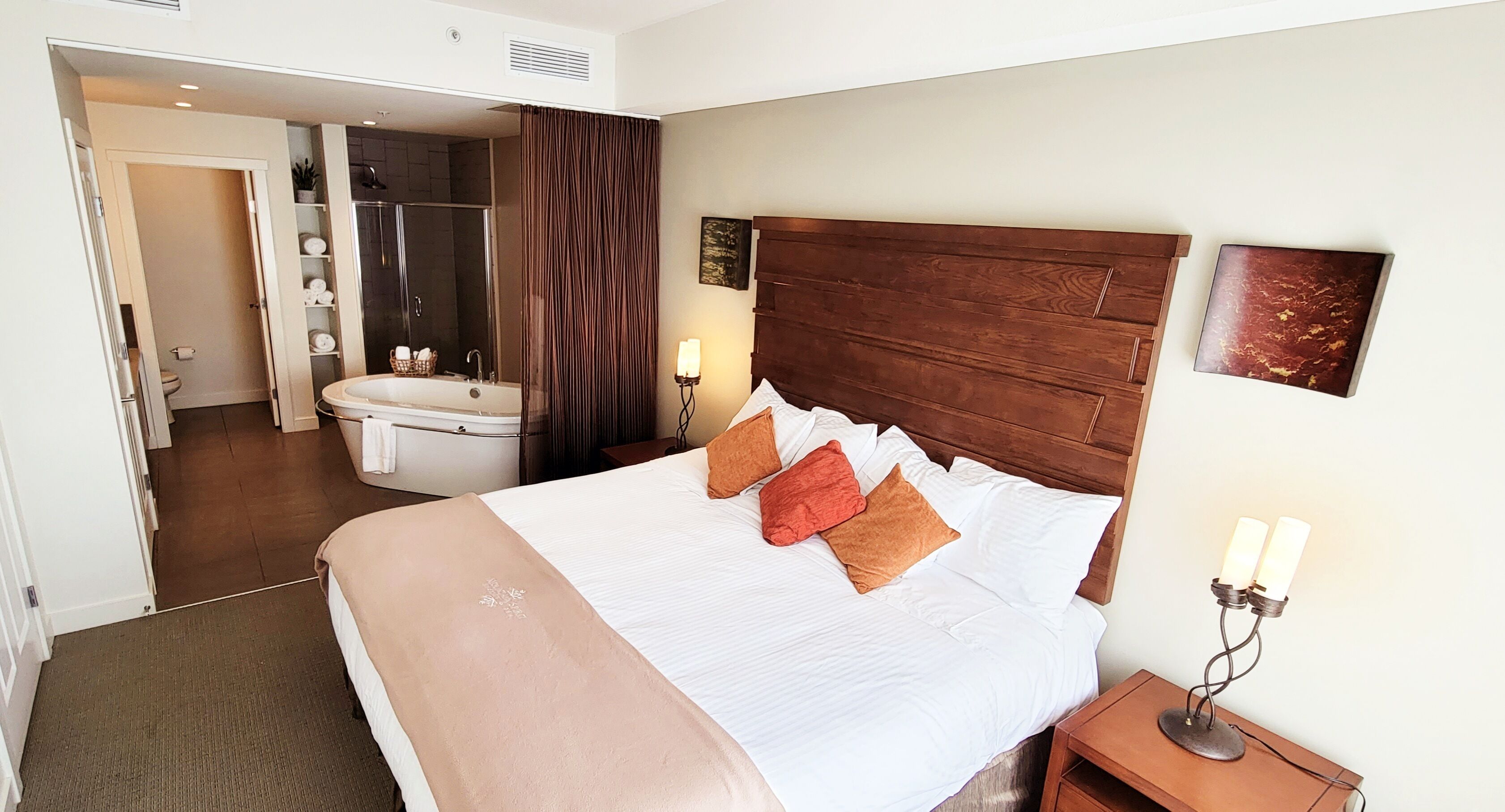 Una habitación de hotel con una cama grande, cabecera de madera, una mesita de noche con lámpara, baño con bañera y ducha.