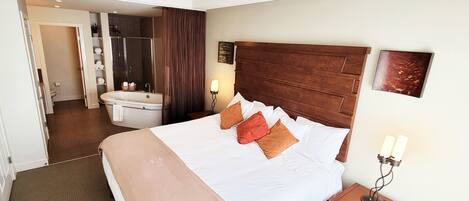 Una habitación de hotel con una cama grande, cabecera de madera, una mesita de noche con lámpara, baño con bañera y ducha.