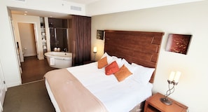 Une chambre d’hôtel avec un grand lit, une tête de lit en bois, une table de chevet avec une lampe, une salle de bain avec une baignoire et une douche.