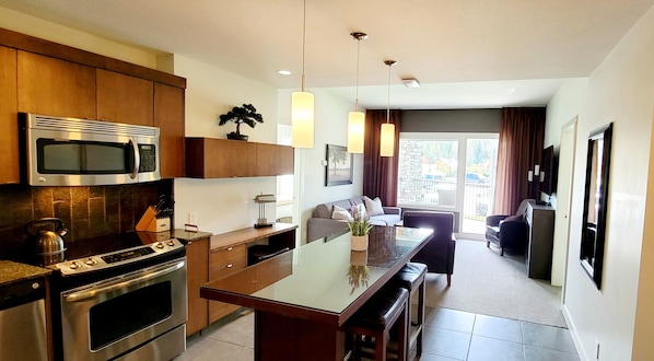 A modern kitchen with stainless steel appliances, a dining area with a glass table, and a living room with a sofa and a TV.