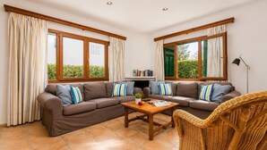 A living room with a sectional sofa, a coffee table, and a wicker chair. There are two windows with curtains, a bookshelf, and a floor lamp. - Villa Binimelis - Five Bedroom Villa, Sleeps 10 (Cala d'Or)