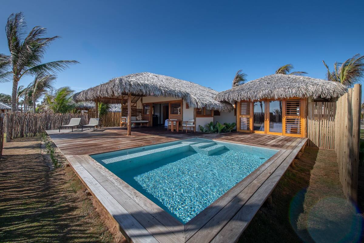 A poolside area with a wooden deck, two thatched-roof structures, and a clear blue pool.