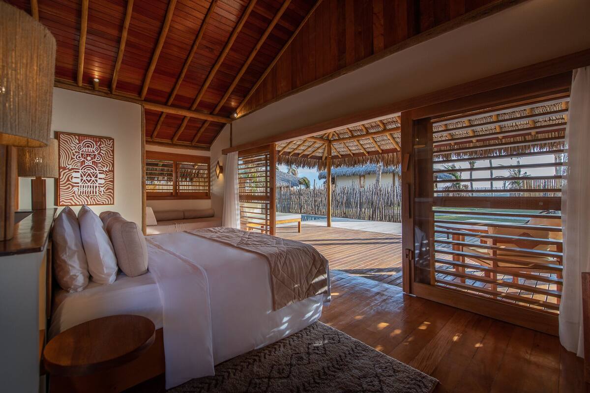 A spacious bedroom with a large bed, wooden ceiling, and a view of an outdoor area with a thatched roof and palm trees.