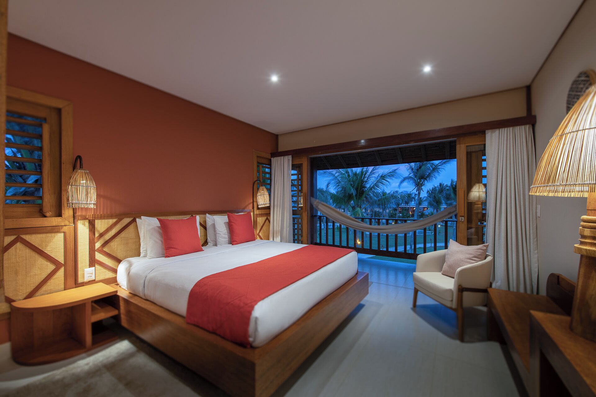 A hotel room with a large bed, a sitting area with a chair, a small table, a balcony with a view of palm trees, and a hammock.