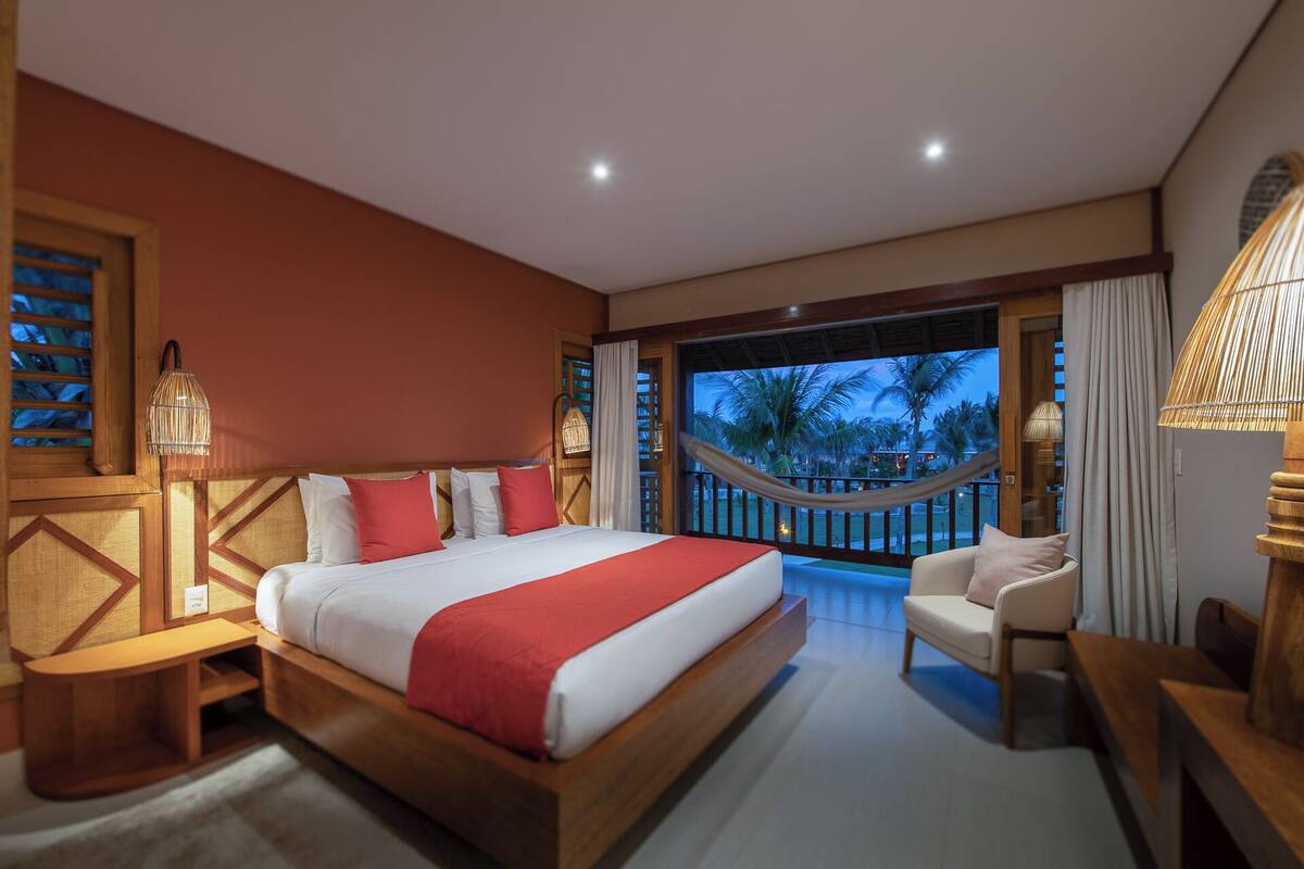 A hotel room with a large bed, a sitting area with a chair, a small table, a balcony with a view of palm trees, and a hammock.