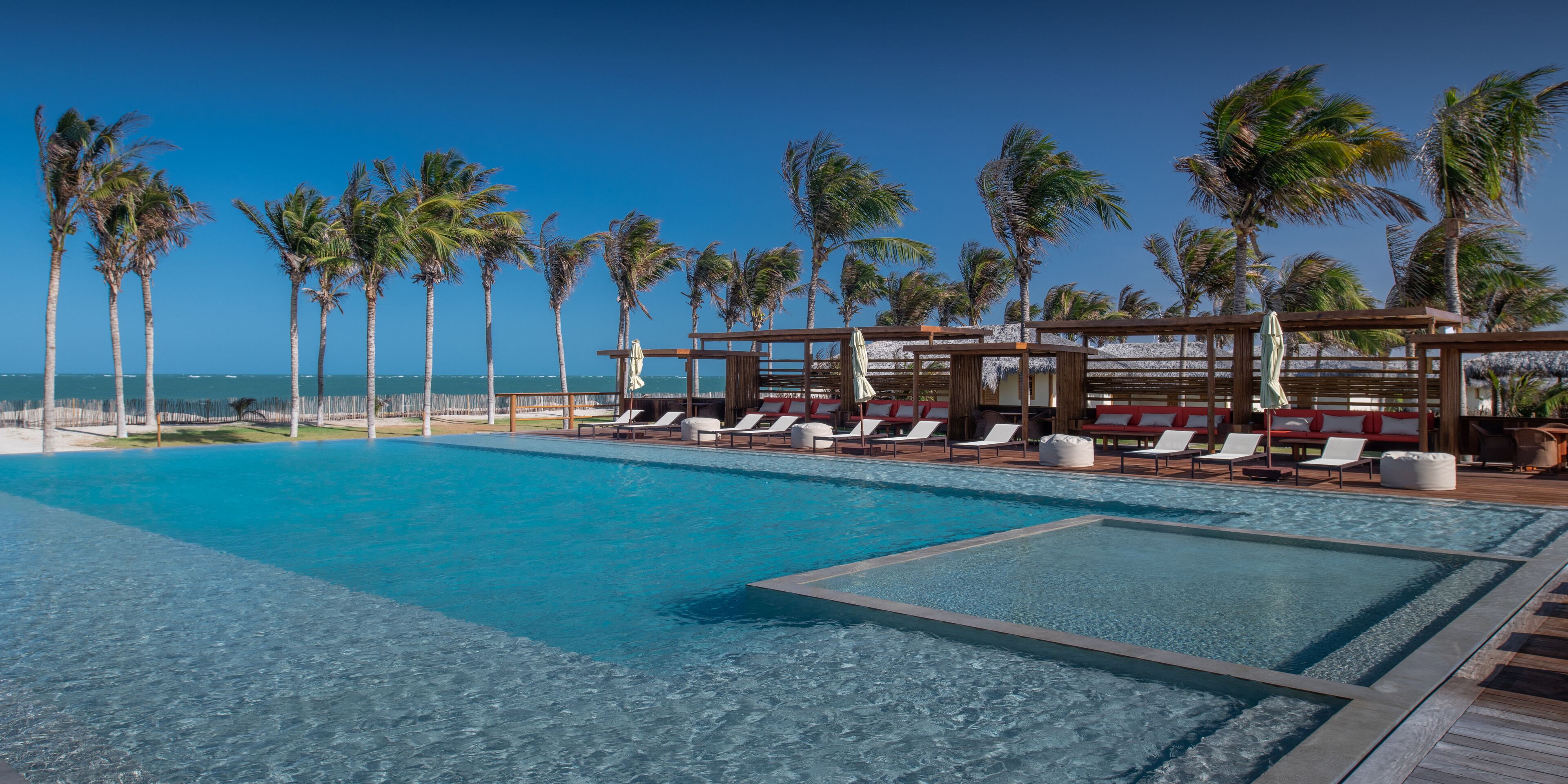 A pool area with lounge chairs and a wooden deck, surrounded by palm trees and a clear blue sky.