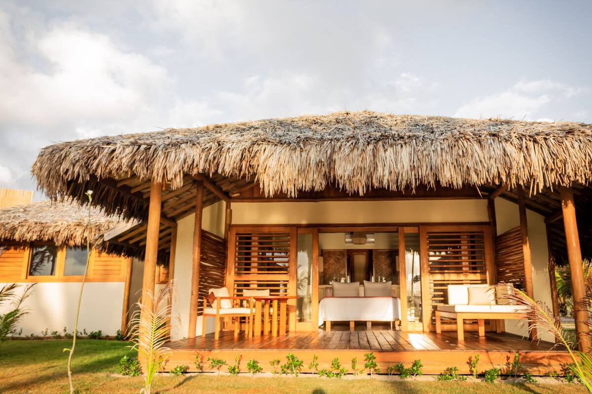 A thatched-roof bungalow with a wooden deck, a bed, and two chairs.