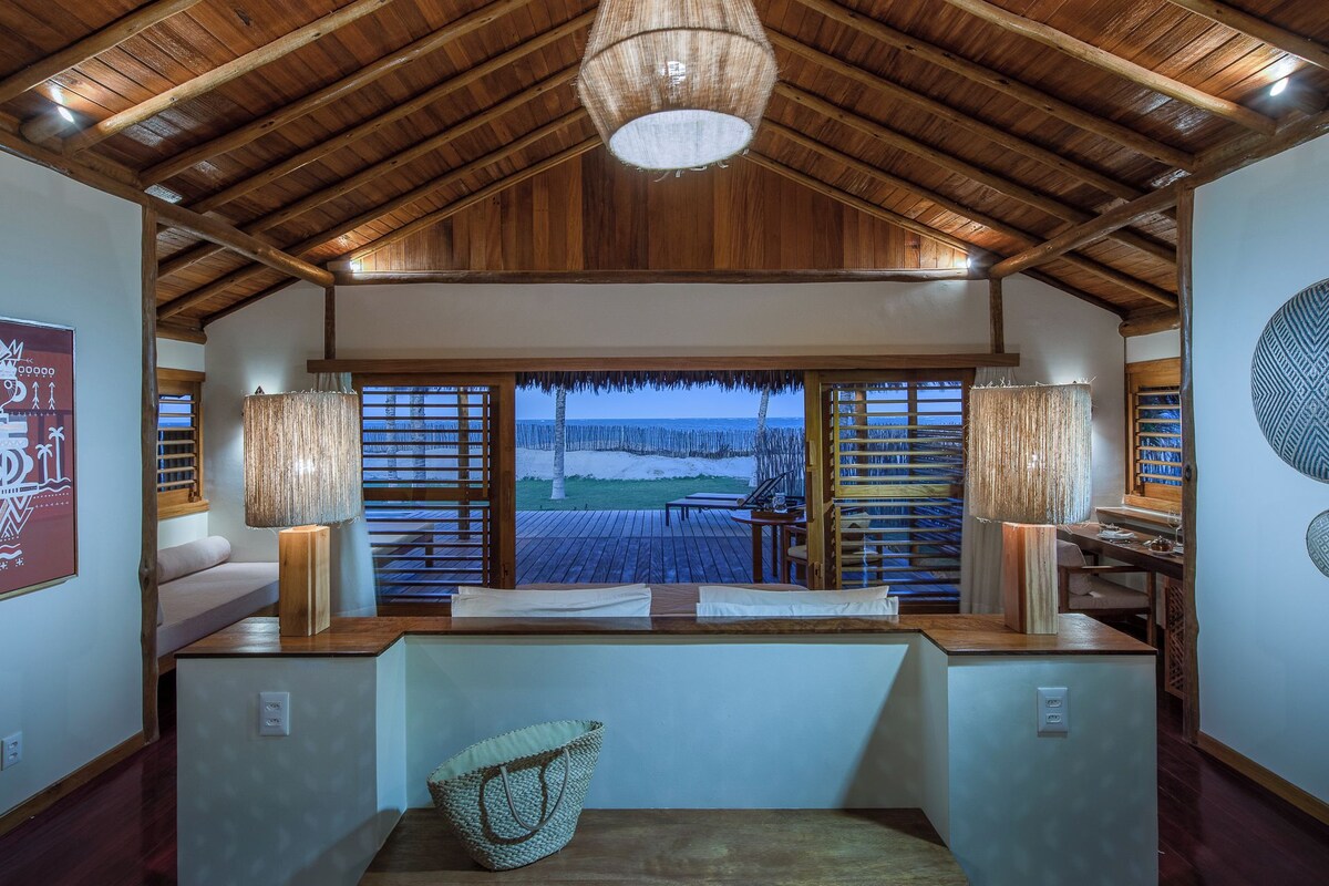 A room with a wooden ceiling, a large window overlooking the beach, a table with chairs, and a hanging light fixture.