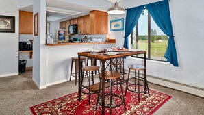 A dining area with a wooden table and four chairs, a red patterned rug, and a kitchen in the background. - Fishy-Fishy Townhome, 2BR/2BA (Kenai)