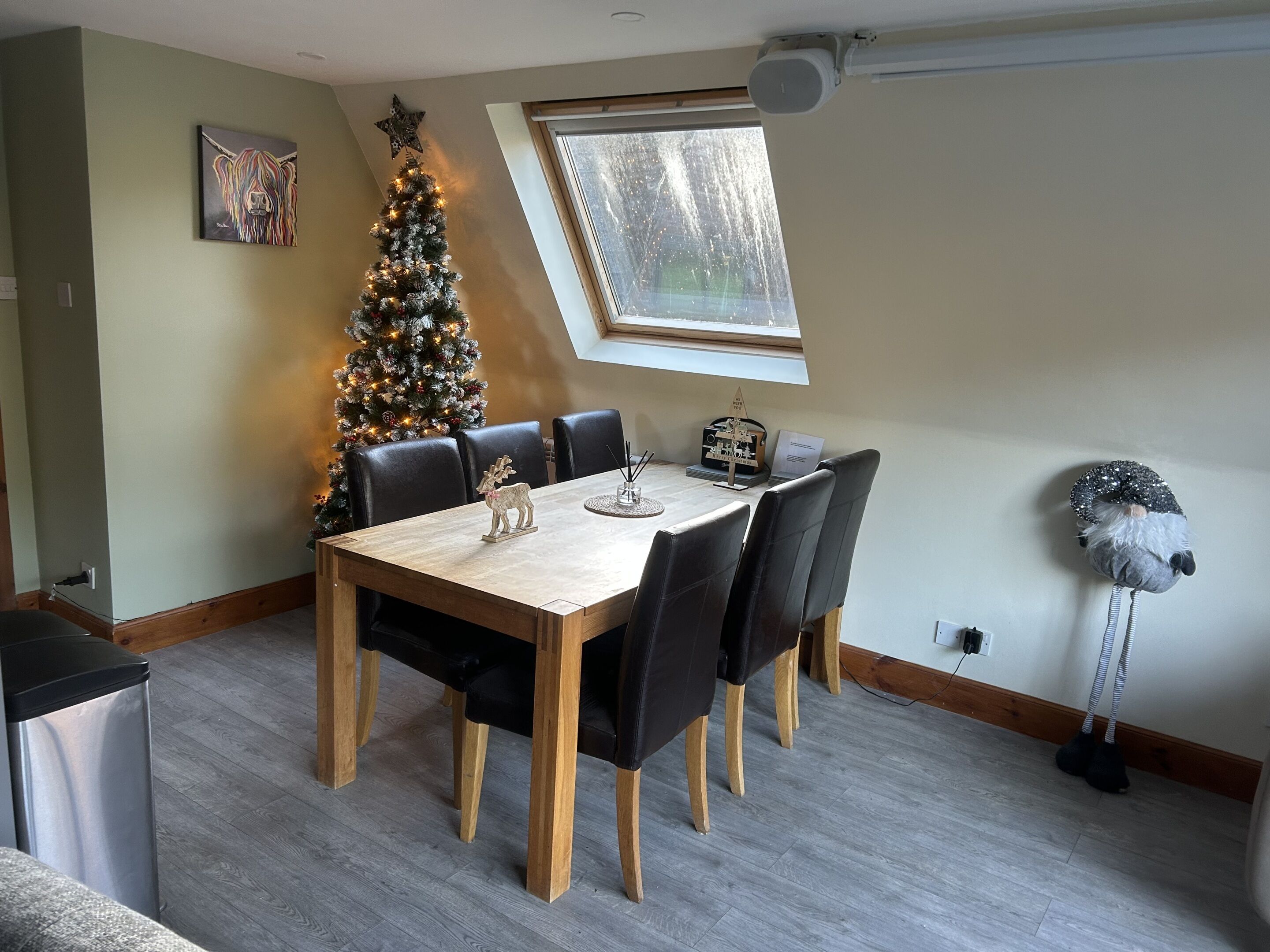 A dining area with a wooden table and black chairs, a Christmas tree, and a window with a view of trees.