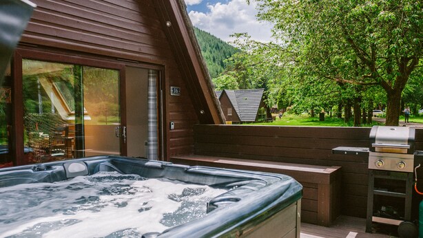 A hot tub with a wooden deck, a barbecue grill, and a view of a forested mountain. - Bright Star Lodge (Invergarry)
