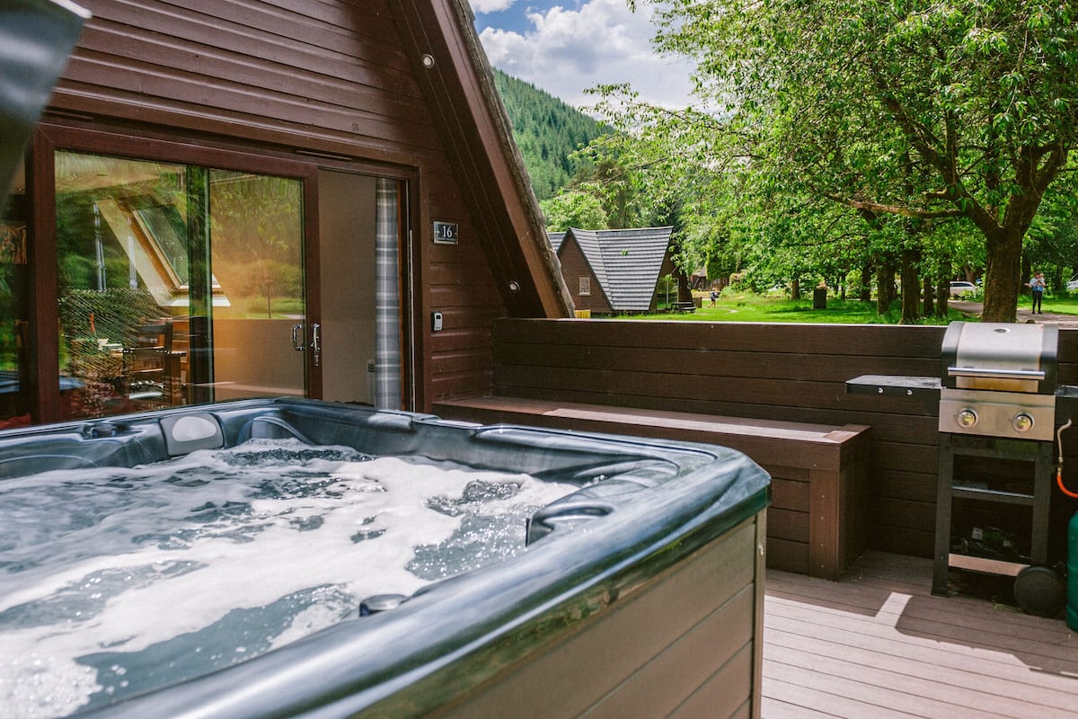 A hot tub with a wooden deck, a barbecue grill, and a view of a forested mountain.