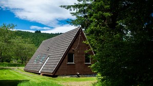 A wooden cabin with a sloped roof, set in a lush green landscape with trees and a hill in the background. - Bright Star Lodge (Invergarry)