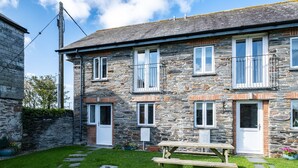 A stone building with multiple windows and doors, a paved pathway, and a wooden bench in the foreground. - Stable Cottage (Port Isaac)