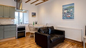 A modern kitchen with wooden beams, a dining area with a white table and chairs, and a black leather sofa. - Stable Cottage (Port Isaac)