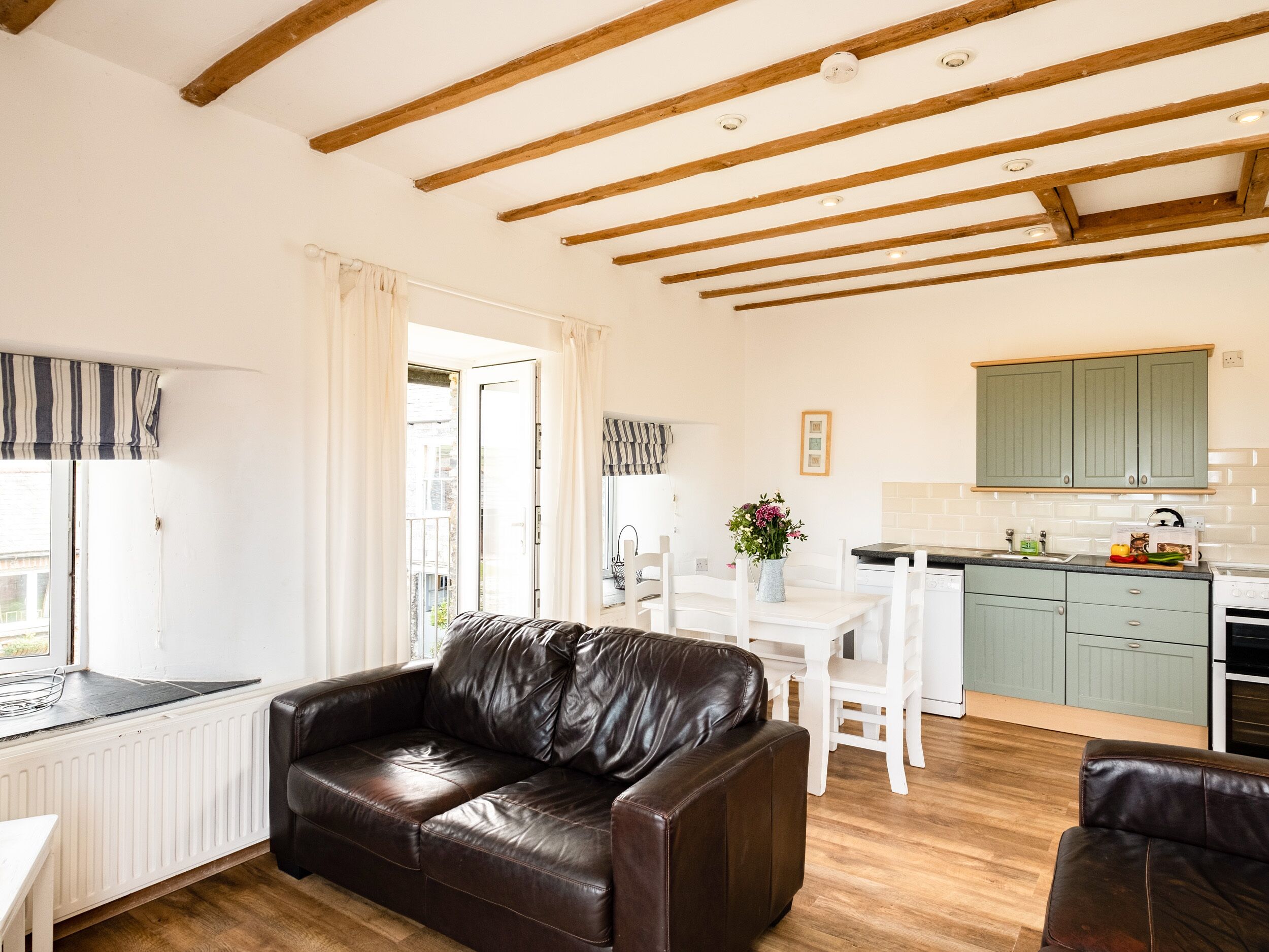 Una sala de estar con un sofá de cuero marrón, una mesa de comedor con sillas, una zona de cocina con armarios verdes y una ventana con cortinas.