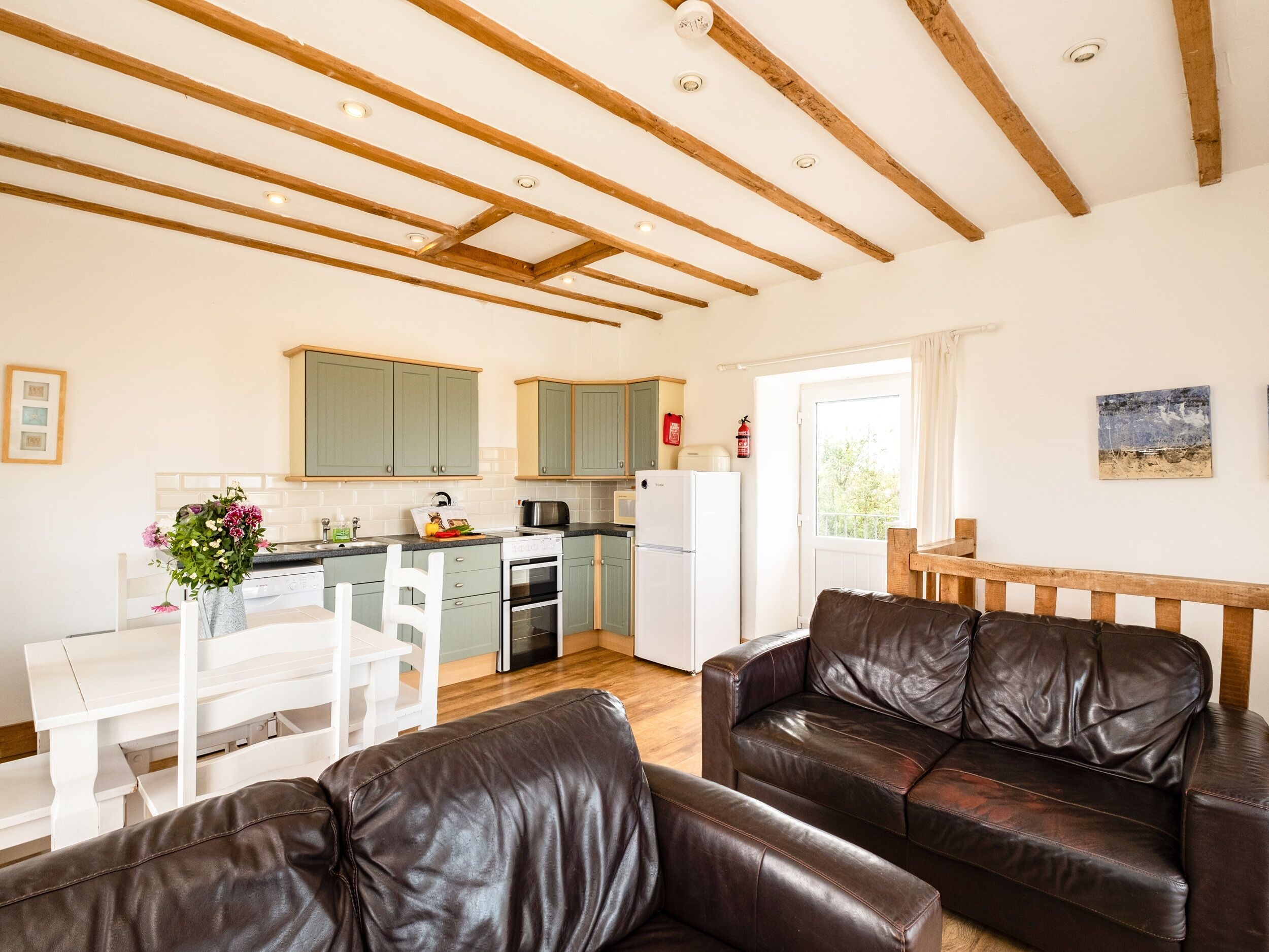 A modern kitchen with wooden beams, a dining area with a white table and chairs, and a leather sofa in a living area.