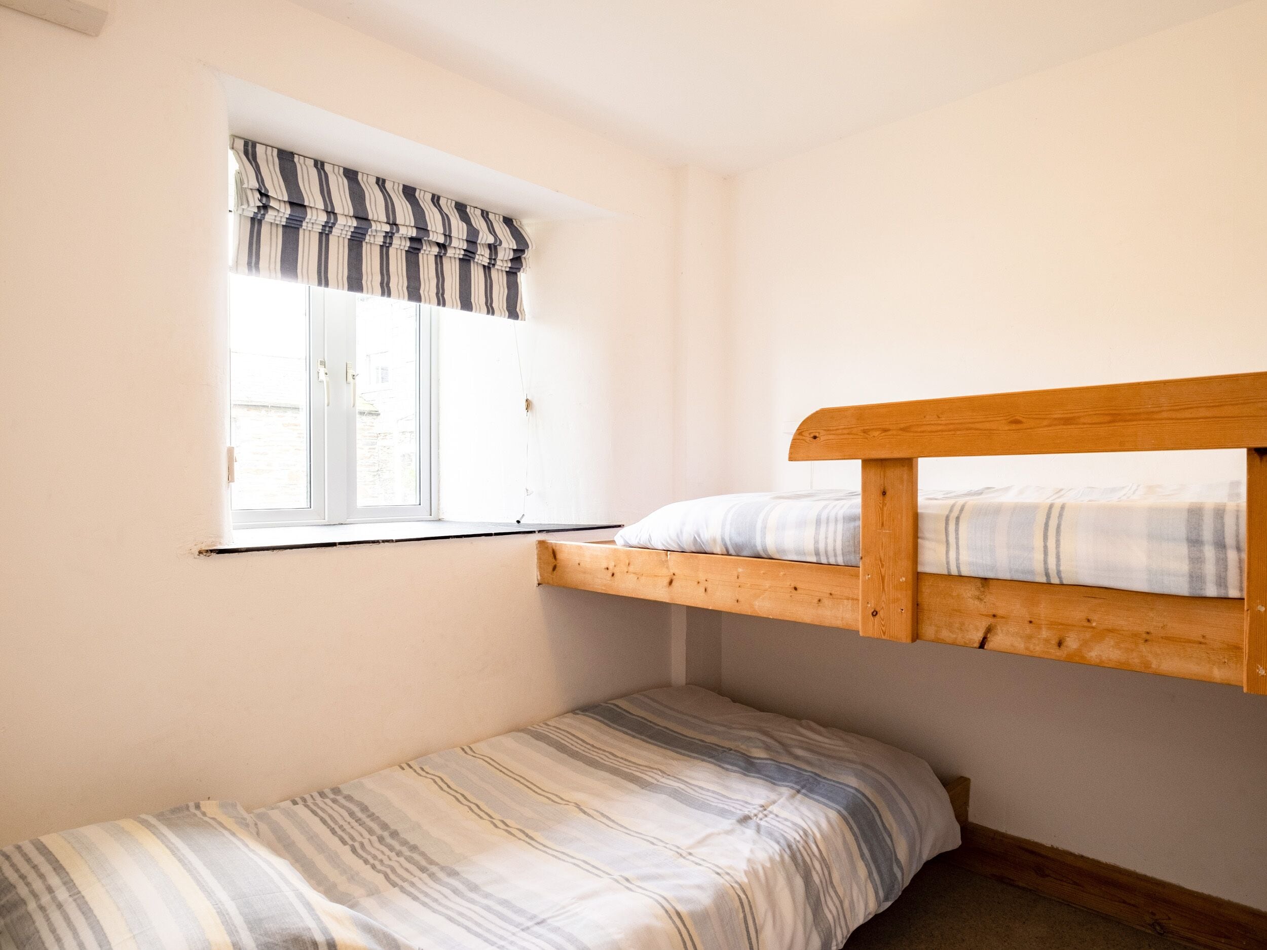 A small, single-room with a bunk bed and a single bed. There is a window with a striped valance and a view of the outside.