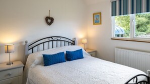 A bedroom with a bed, two bedside tables, a lamp, a window with a view of greenery, a heart-shaped decoration, and a framed picture on the wall. - Atlantic Lodge (Port Isaac)