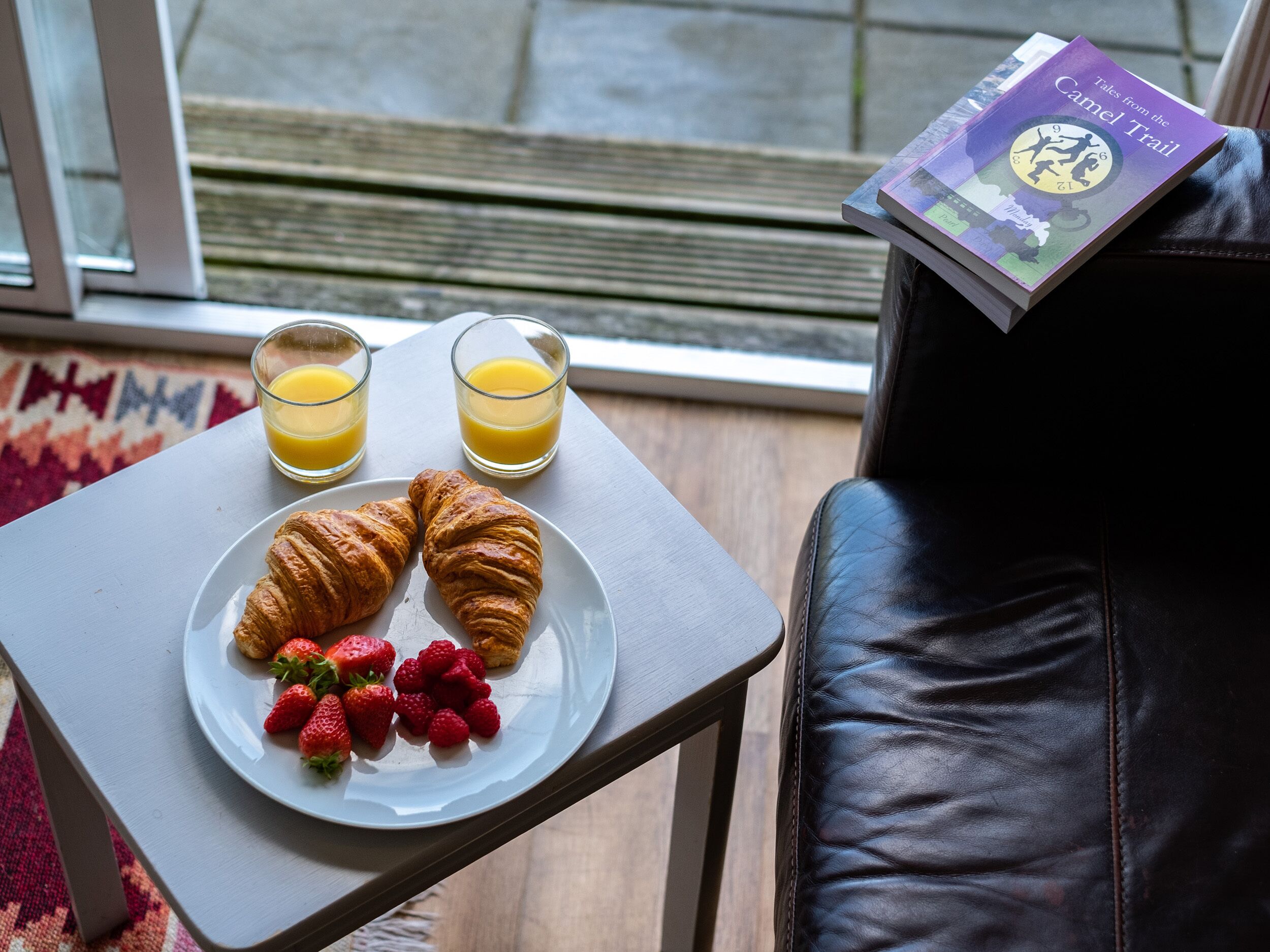 Une table mise pour le petit-déjeuner avec des croissants, des fraises et du jus d’orange, et un livre intitulé « Contes du sentier du chameau » posé sur une chaise.