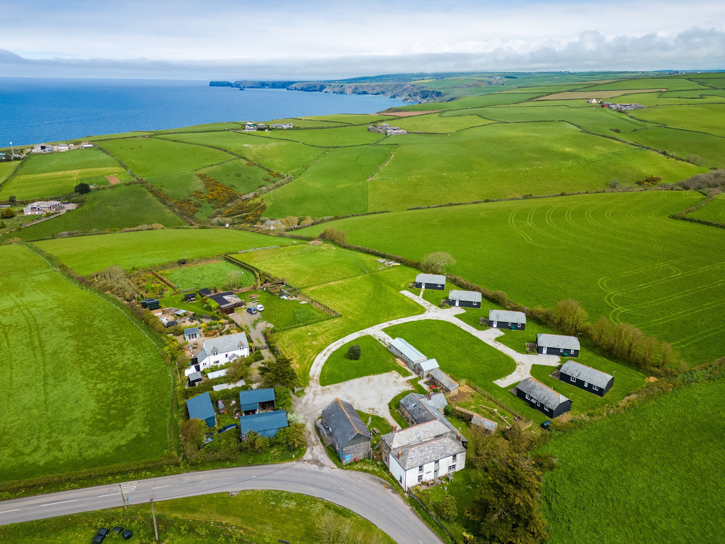 An aerial view of a rural area with green fields, a winding road, and several buildings including houses and a farm structure.