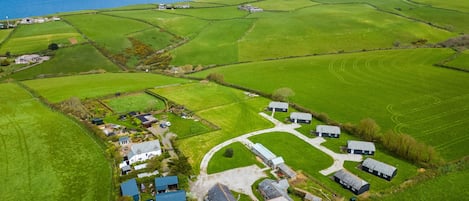 Oversigtsbillede fra luften af et landligt område med grønne marker, en snoet vej og flere bygninger, herunder huse og en landbrugsbygning.