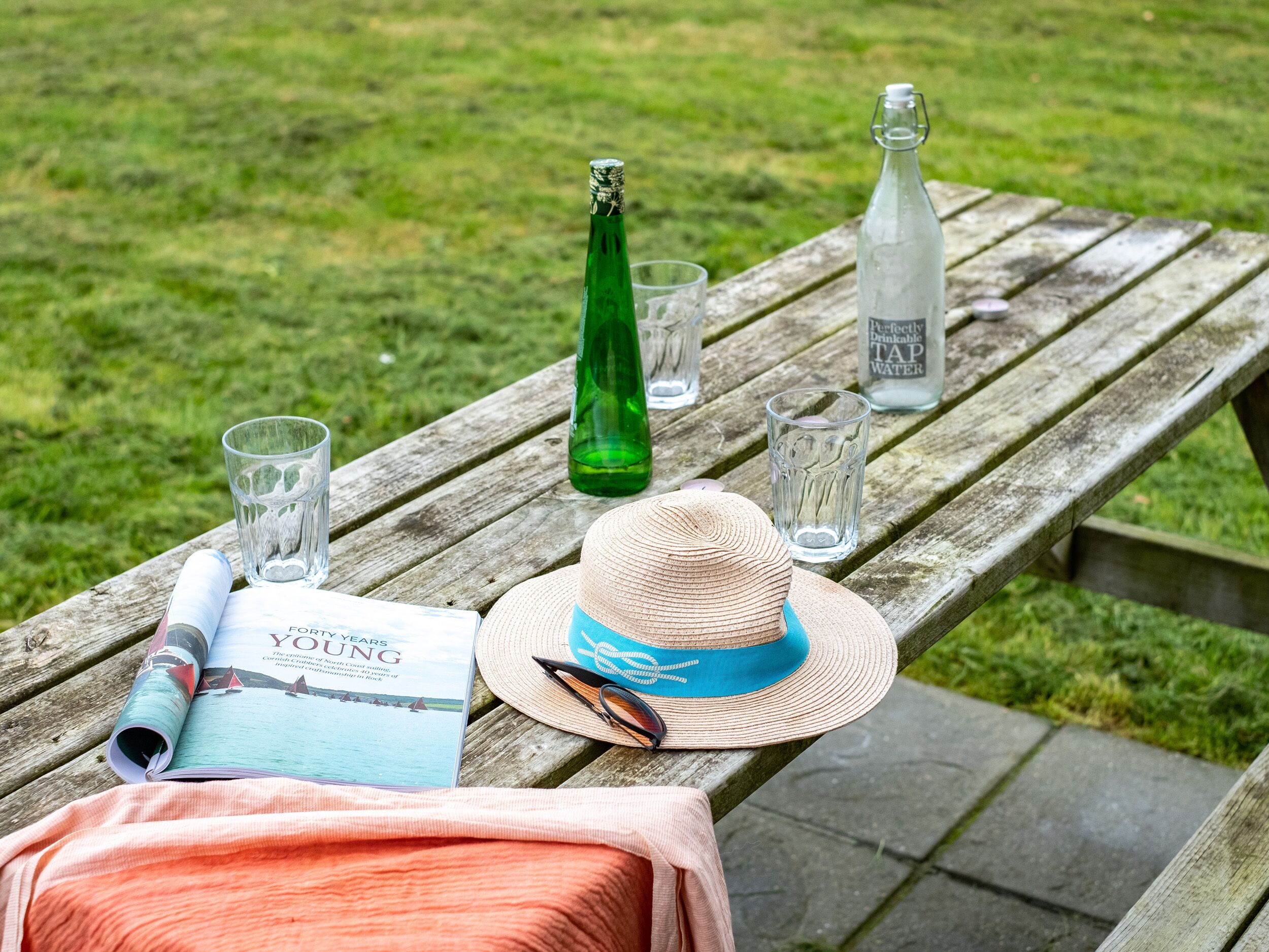 A wooden picnic table with a book, a straw hat, a bottle, glasses, and a water bottle.