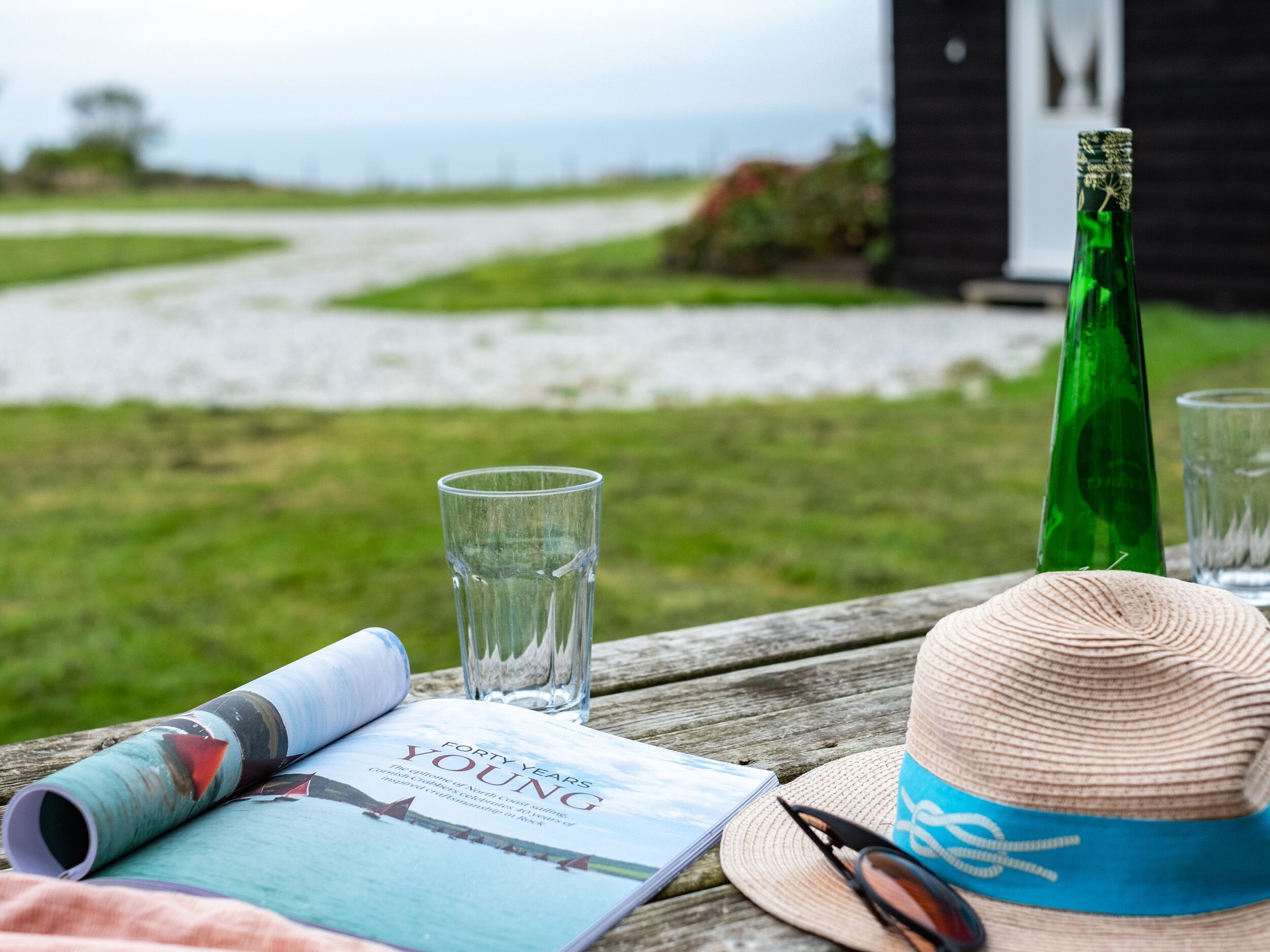 Een houten tafel met een boek, brillen, een strohoed en een groene fles.