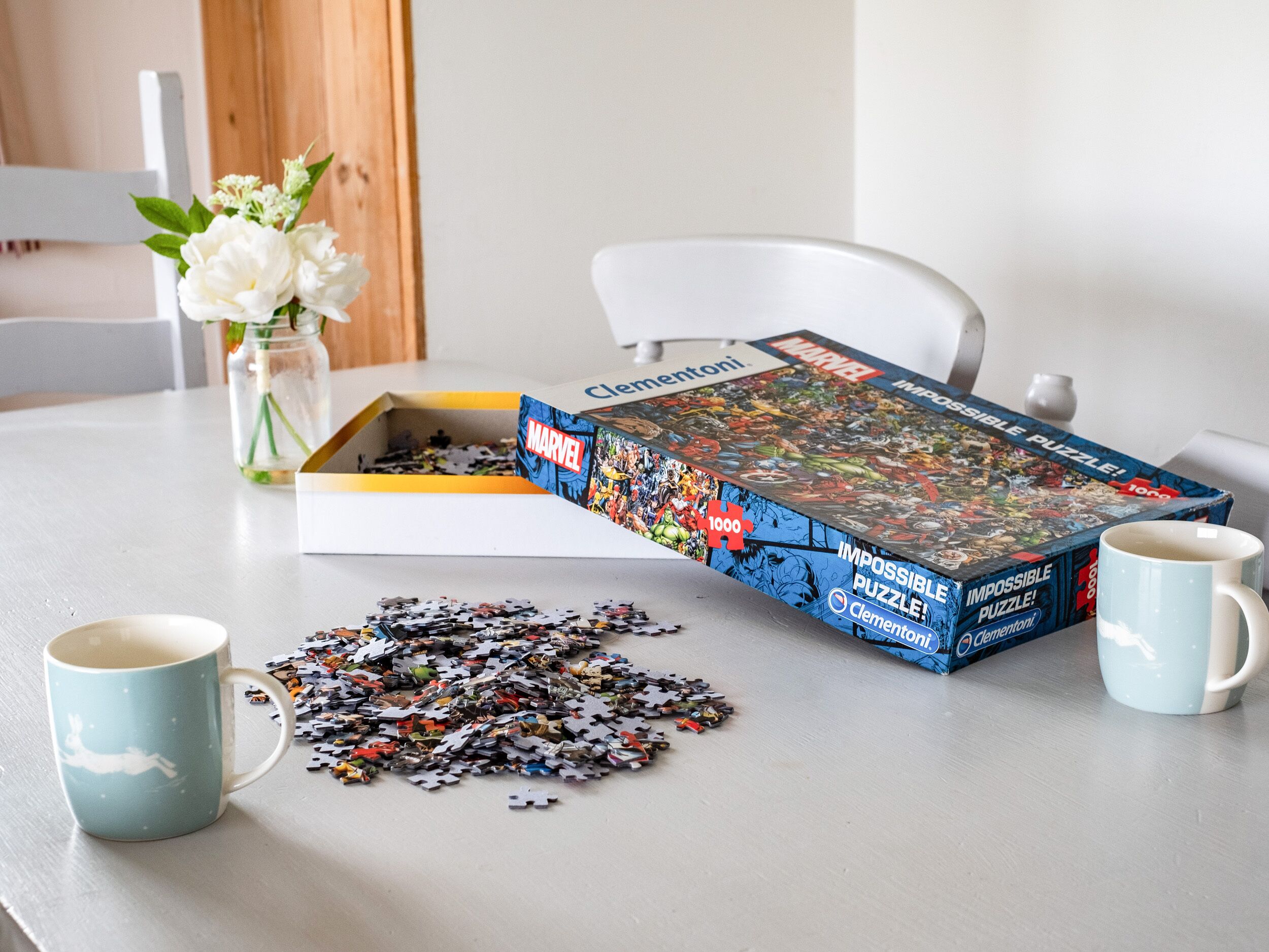 A puzzle box labeled "Marvel" on a table with a partially completed puzzle, a mug, and a vase with flowers.