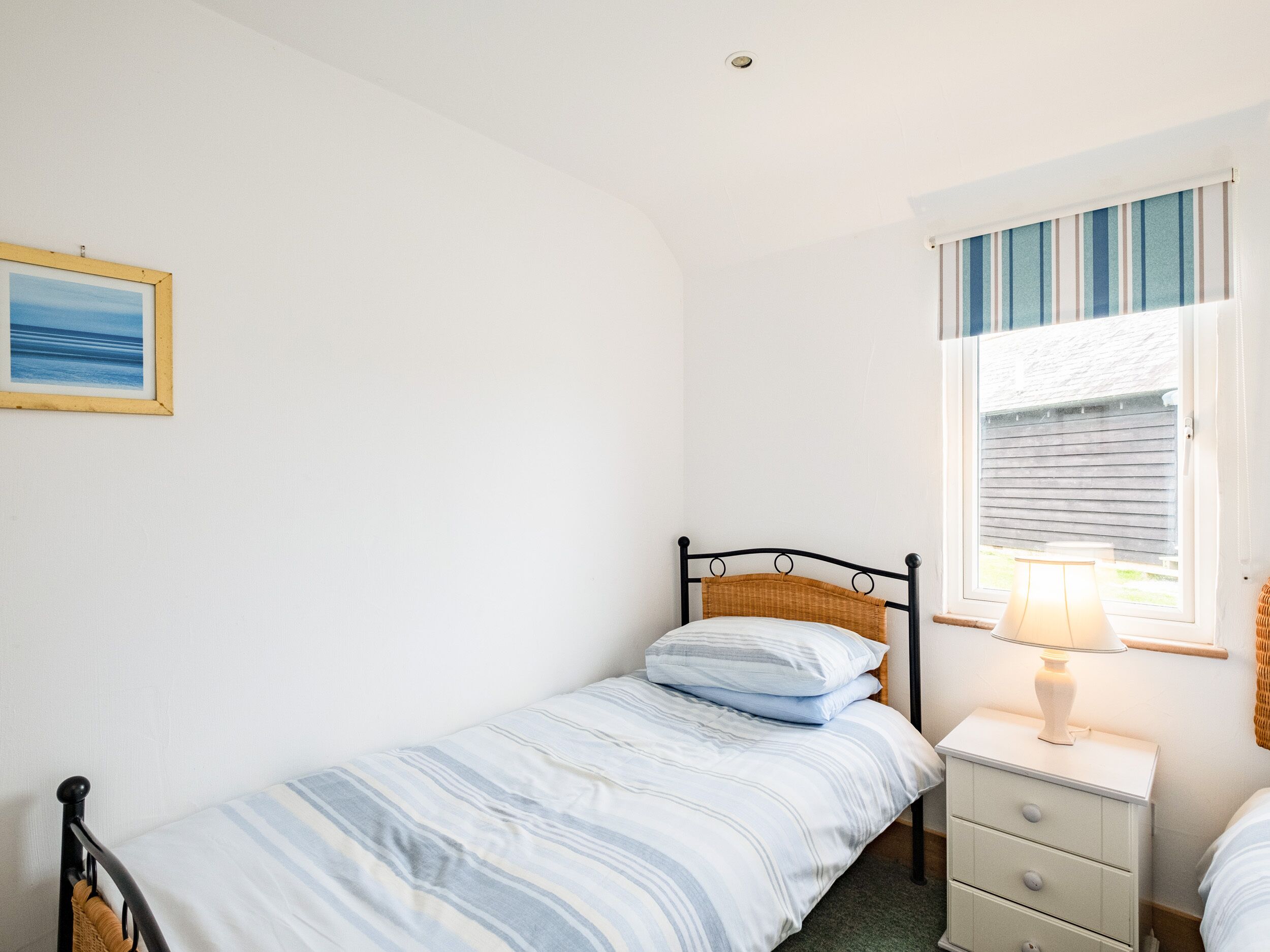 A small, single-bed room with a plain white wall, a framed picture, a window with blinds, and a bedside table with a lamp.