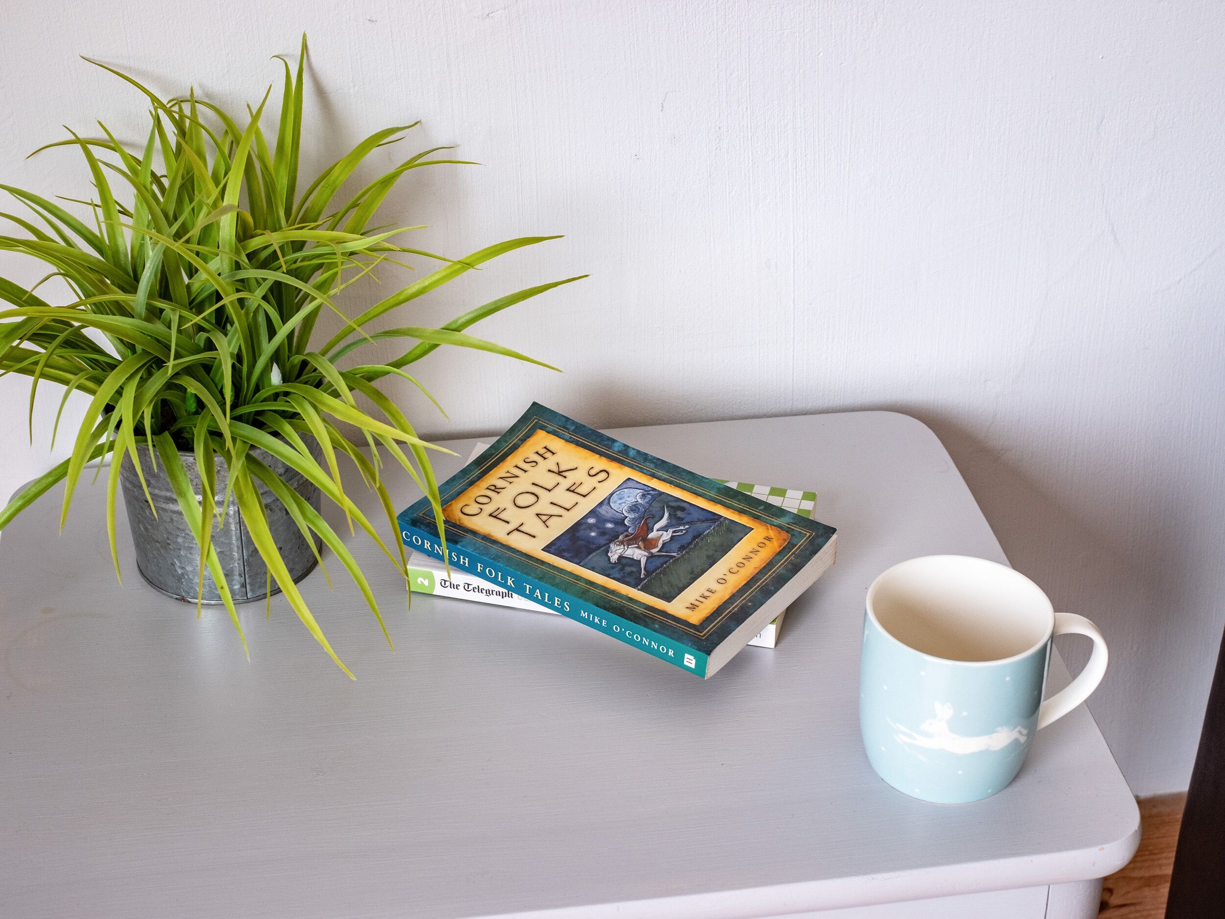 A book titled "Cornish Folk Tales" on a table with a potted plant and a mug.