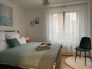 A neatly made bed with pillows, a bedside lamp, and a chair in a room with a large window and sheer curtains.