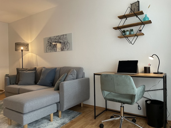 A modern living room with a grey sectional sofa, a desk with a laptop, a standing desk lamp, and a wall-mounted shelf with plants.