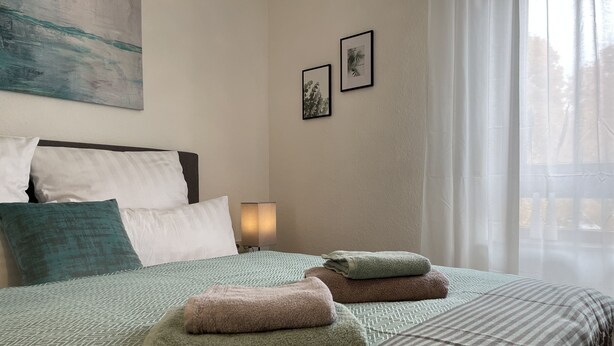 Een net opgemaakt bed met een beddekleed met patroon, kussens en opgevouwen handdoeken. Er staat een bedlampje en een ingelijst beeld op de muur.